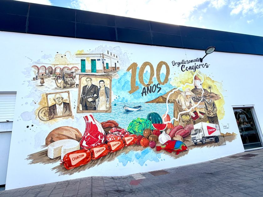 La conocida firma de alimentación celebra un siglo de arraigo social y económico en la isla de los volcanes. En este sentido, la fachada de sus instalaciones principales luce una espectacular obra pictórica inspirada en sus orígenes.
Por: Redacción Mass Cultura | 9 de junio de 2026
(Línea de separación)
El panorama urbano de Arrecife suma una nueva e importante intervención artística de gran formato. Por este motivo, la dirección empresarial ha presentado la pieza realizada para conmemorar oficialmente sus cien años de andadura. La obra decora el exterior del edificio corporativo del barrio portuario. Si buscas conocer el nuevo mural candido armas chacon valterra, aquí te desglosamos todos sus detalles históricos y técnicos.
🎨 De la Recova a Valterra: el homenaje artístico a la trayectoria de la familia Chacón
Por lo que respecta a las dimensiones del proyecto, la pintura cuenta con once metros de ancho por seis de alto. Para su ejecución, el creador se basó en fotografías antiguas facilitadas por los propios familiares:
Escenas tradicionales: El lienzo exterior plasma con mimo las labores de la agricultura y la pesca tradicional conejera.
El autor: La pieza está firmada por Iván T., un reconocido artista argentino afincado en tierras lanzaroteñas.
Declaraciones del artista: "Es un honor transmitir con mi arte parte de la historia de una empresa tan querida", señaló el pintor.
🥩 El legado de Cándido Armas Chacón y el producto local en una obra monumental
Por otra parte, la evolución comercial de la marca refleja el crecimiento de la propia sociedad insular. El bisnieto del fundador, Carlos, recordó que los primeros pasos se dieron tras el traslado familiar desde la vecina isla de Fuerteventura. De esta manera, las recetas tradicionales de Cándido Armas Chacón pasaron de un pequeño puesto en la Recova a una carnicería en la calle Fajardo.
Como resultado de este siglo de esfuerzo, la marca se ha consolidado como una referencia absoluta en el sector alimentario. Asimismo, el chorizo de la casa destaca por ser el único embutido tradicional elaborado íntegramente en la isla. En definitiva, la pintura exterior funcionará como un tributo permanente al desarrollo económico de la capital. De igual forma, a través de las secciones de arte urbano en Mass Cultura seguiremos poniendo en valor la transformación visual de nuestros barrios.